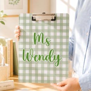 May include: A rectangular clipboard featuring a light sage green and white gingham check pattern. The text 'Ms Wendy' is printed in a dark green cursive font in the center. A silver metal clip is attached to the top edge of the clipboard.