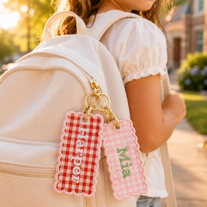 Peut inclure: Deux étiquettes de sac rectangulaires à bords festonnés attachées à un sac à dos crème avec des anneaux en métal doré. Une étiquette présente un motif vichy rouge et blanc avec le nom Harper brodé en blanc, et l'autre est vichy rose clair avec le nom Mia brodé en vert.