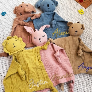 May include: A collection of soft, plush baby security blankets in various animal shapes, including an elephant, koala, bear, lion, and rabbit. Each blanket is a different color and has a name embroidered on it. The blankets are made of a textured fabric.
