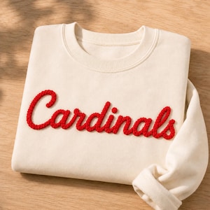 Może przedstawiać: Kremowa bluza z okrągłym dekoltem i napisem Cardinals wyhaftowanym wypukłą, jaskrawoczerwoną czcionką kursywą na piersi. Odzież posiada długie rękawy oraz prążkowany kołnierz, co nadaje jej prosty i schludny wygląd.