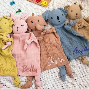 Puede incluir: Una colección de mantas de seguridad para bebés con forma de animales de peluche suave en varios colores, incluyendo amarillo, rosa, marrón, azul y beige. Cada manta presenta una cara de animal diferente y un nombre bordado.