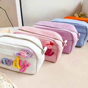 May include: A row of five rectangular corduroy pouches in white, pink, lavender, light blue, and orange. Each pouch features a metal zipper and a personalized name in colorful crocheted yarn, accented with small crocheted flowers or butterflies.