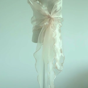 May include: A sheer ivory shawl with a scalloped edge and gold trim. The shawl is draped over a dress form.
