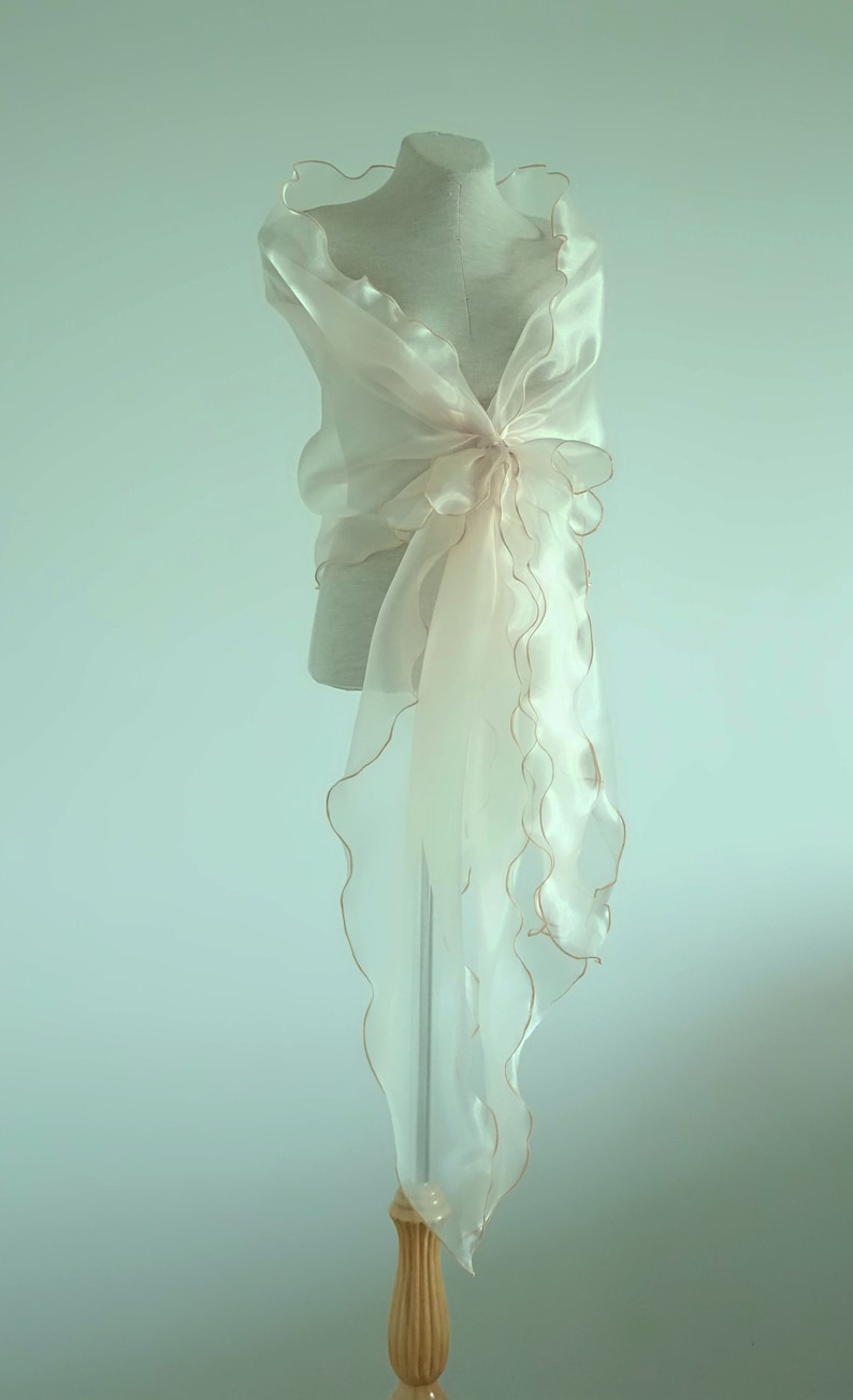 May include: A sheer ivory shawl with a scalloped edge and gold trim. The shawl is draped over a dress form.