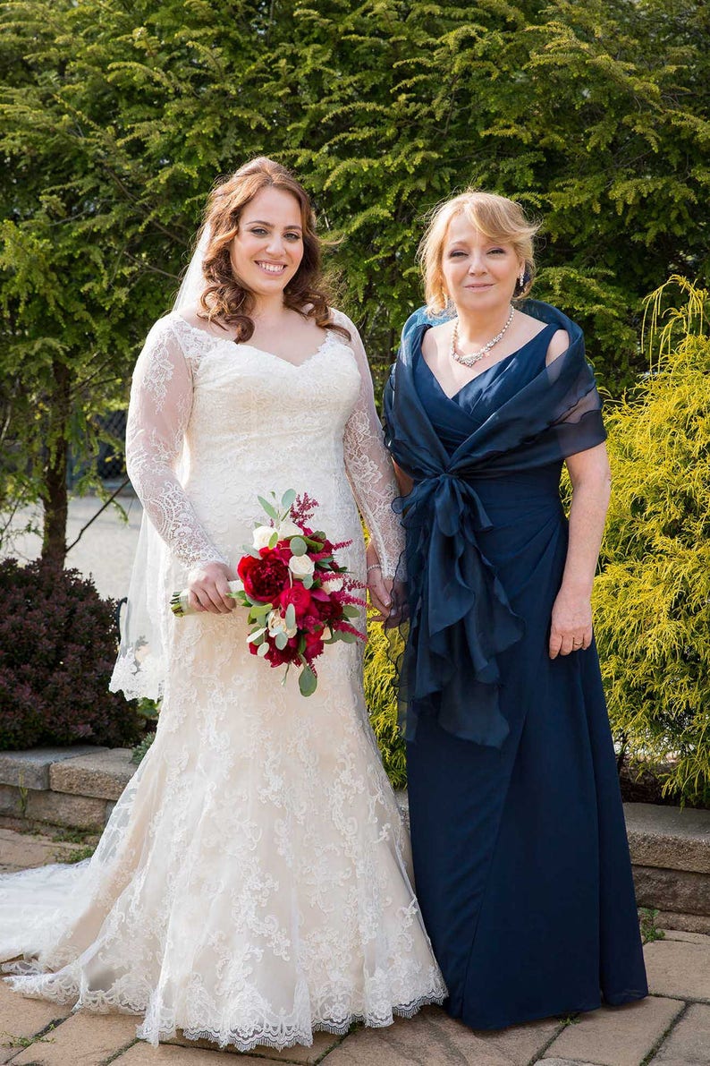 K&ouml;nnte beinhalten: Eine Braut in einem wei&szlig;en Spitzen-Hochzeitskleid mit langem Schleier h&auml;lt einen Strau&szlig; roter und wei&szlig;er Blumen. Sie steht neben ihrer Mutter, die ein marineblaues Kleid mit passendem Schal tr&auml;gt.