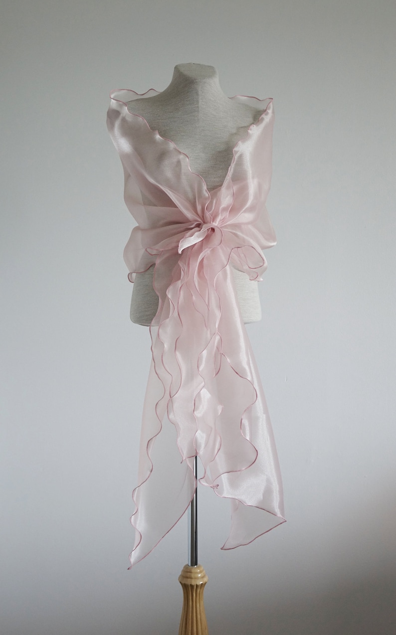 May include: A pale pink sheer shawl with ruffled edges, draped over a mannequin. The shawl is tied in a bow at the front.