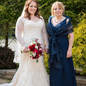 May include: A bride in a white lace wedding dress holds a bouquet of red and white flowers. She stands next to a woman in a navy blue dress with a matching shawl.
