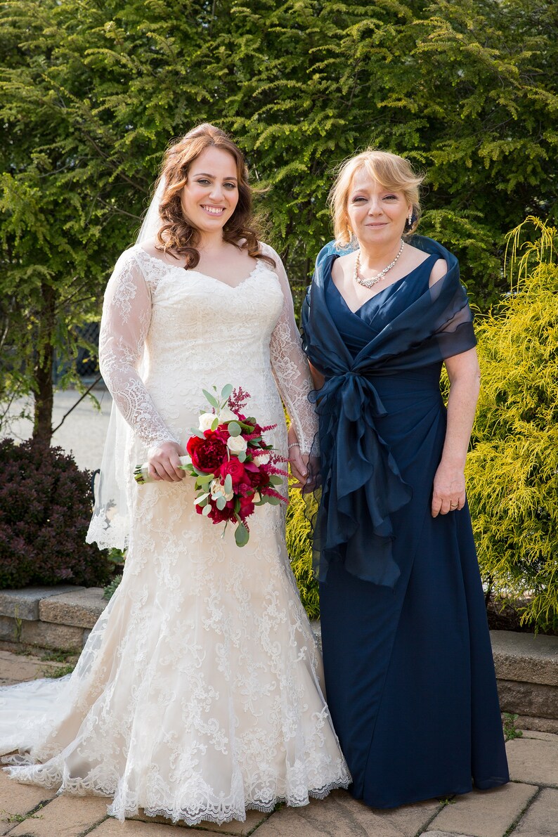 May include: A bride in a white lace wedding dress holds a bouquet of red and white flowers. She stands next to a woman in a navy blue dress with a matching shawl.