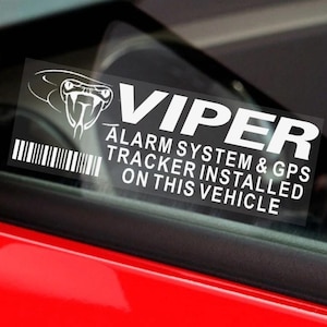 May include: A white vinyl sticker with the word "VIPER" in bold letters, a snake head graphic, and the text "ALARM SYSTEM & GPS TRACKER INSTALLED ON THIS VEHICLE." The sticker is affixed to a car window.