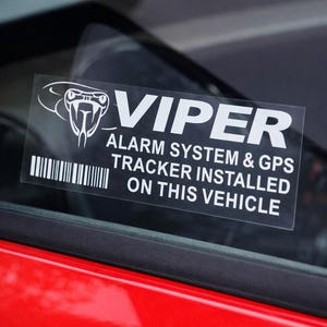 May include: A clear sticker on a car window with the word "VIPER" in large white letters. Below, text reads "ALARM SYSTEM & GPS TRACKER INSTALLED ON THIS VEHICLE." A white snake head logo is above the word "VIPER".