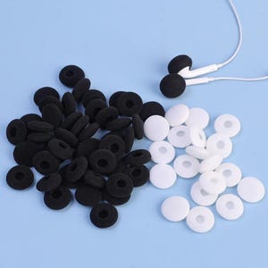 Pode incluir: Uma coleção de capas de fone de ouvido pretas e brancas e um par de fones de ouvido brancos com capas pretas, tudo em um fundo azul claro. As capas de fone de ouvido são feitas de espuma e estão em vários tamanhos.