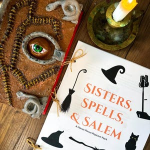 Może przedstawiać: Brązowa, teksturowana książka z dużym okiem i detalami przypominającymi węża, obok białej kartki z napisem "Sisters, Spells, & Salem". Obecna jest również zapalona świeca w mosiężnym świeczniku.