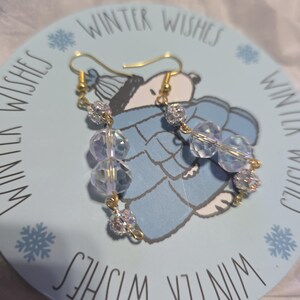 May include: A pair of dangle earrings with clear, faceted beads and small, sparkly silver accents. The earrings have gold-colored hooks and are displayed on a light blue decorative plate with the words "Winter Wishes".