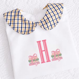 May include: White baby romper with a Peter Pan collar in a yellow, navy, and white check pattern. The romper features a pink embroidered initial "H" and stacks of books with pink bows.