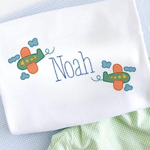 Op de afbeelding: Witte babydeken geborduurd met de naam "Noah" in blauw schrift. Twee groene en oranje vliegtuigen met blauwe wolken zijn op de deken geborduurd. De deken ligt op een blauw-wit geruit oppervlak.