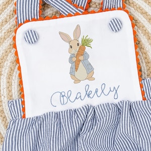 Könnte beinhalten: Weiße Baby-Latzhose mit blau-weiß gestreiftem Stoff und orangefarbenem Besatz. Das Lätzchen zeigt ein gesticktes Bild eines Hasen mit einer Karotte und den Namen "Blakely" darunter.