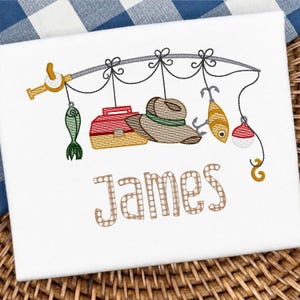 May include: White fabric featuring an embroidered fishing design. The design includes a fishing rod, tackle box, hat, fish, and the name "James" in a decorative font. The colour palette includes shades of brown, red, green, and yellow.