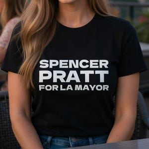 May include: A black t-shirt with the text "SPENCER PRATT FOR LA MAYOR" in white block letters. The image shows a person wearing the shirt, smiling, with a city skyline in the background. The shirt is a classic crew neck style.
