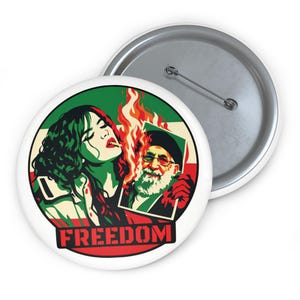 Puede incluir: Botón blanco con un diseño gráfico que presenta a una mujer con piel verde y una imagen de un hombre en llamas. La palabra "FREEDOM" está en texto rojo en la parte inferior. El botón tiene un respaldo de alfiler plateado.