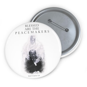 Puede incluir: Botón blanco con un alfiler plateado. El botón presenta una imagen en blanco y negro de un hombre con una figura detrás. El texto "BLESSED ARE THE PEACEMAKERS" y "MATTHEW 5:9" están impresos en el botón.
