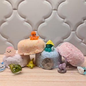 Könnte beinhalten: Mehrere runde, gesprenkelte Badebomben in Pfirsich-, Blau- und Rosatönen, begleitet von kleinen, bunten Kunststofffiguren von Meerestieren. Eine 25-Cent-Münze liegt zur Größenreferenz an der blauen Badebombe.