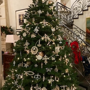 May include: A festive Christmas tree adorned with a variety of ornaments and fairy lights. The decorations include crosses, stars, and other shapes. The tree is in a well-lit room, with a staircase visible in the background.
