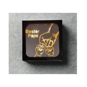 Puede incluir: Luz de noche cuadrada con marco de madera. El interior iluminado muestra una ilustración dorada de una mano sosteniendo dos puños más pequeños. El texto "Bester Papa" y los nombres "Maila" y "Juna" también son dorados.