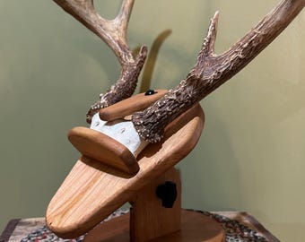 May include: A wooden deer antler mount with a natural finish. The mount features a light brown wooden base and a curved platform to hold the antlers. The antlers are a light brown color with a textured surface.