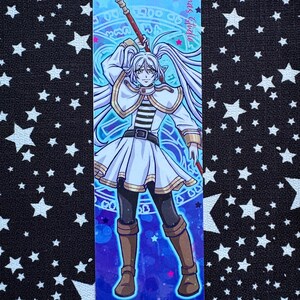May include: A bookmark with an anime-style illustration of a character with long white hair, wearing a white and gold outfit, and holding a staff. The background is a gradient of blues and purples with magical symbols. Text on the bookmark reads "Artimas Studio".