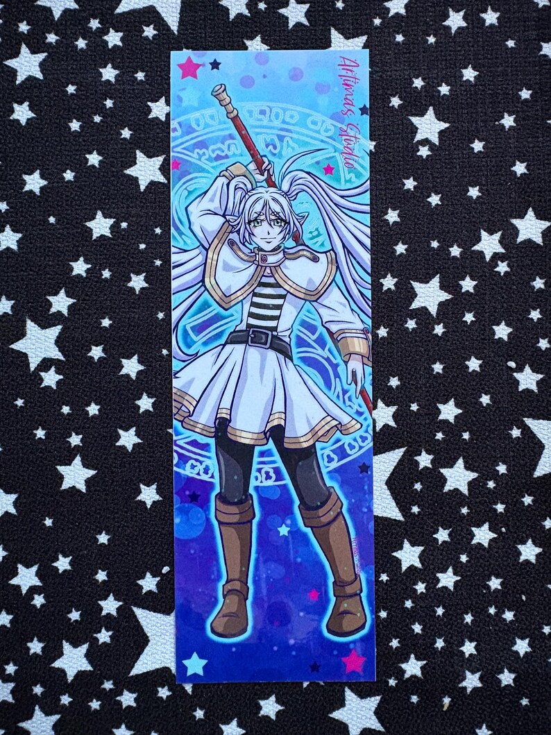 May include: A bookmark with an anime-style illustration of a character with long white hair, wearing a white and gold outfit, and holding a staff. The background is a gradient of blues and purples with magical symbols. Text on the bookmark reads "Artimas Studio".