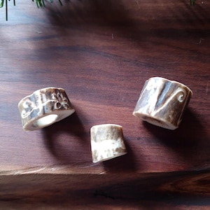 May include: Three antler rings, each with a unique pattern and natural color variations of brown and cream. The rings are displayed on a dark wood surface, showcasing their rustic charm and organic texture.