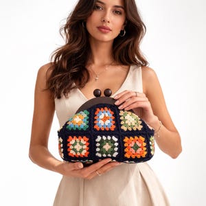 May include: A navy blue crochet clutch with a wooden clasp. The clutch features a colorful patchwork design with squares of orange, green, blue, and cream. The bag is held by a person.