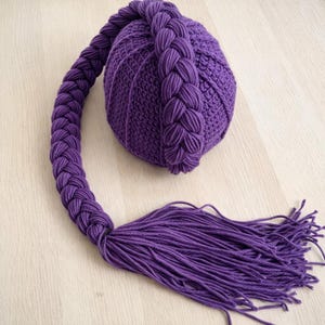 May include: A handcrafted, purple crocheted bag with a braided strap and a tassel. The bag is round with vertical ribbing. The strap is a thick braid, and the tassel is made of many strands of yarn.