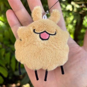 May include: A tan-coloured plush keychain in the shape of a cartoon animal. It has small, upright ears, a smiling face with a pink tongue, and three black legs. The keychain is suspended by a black string.