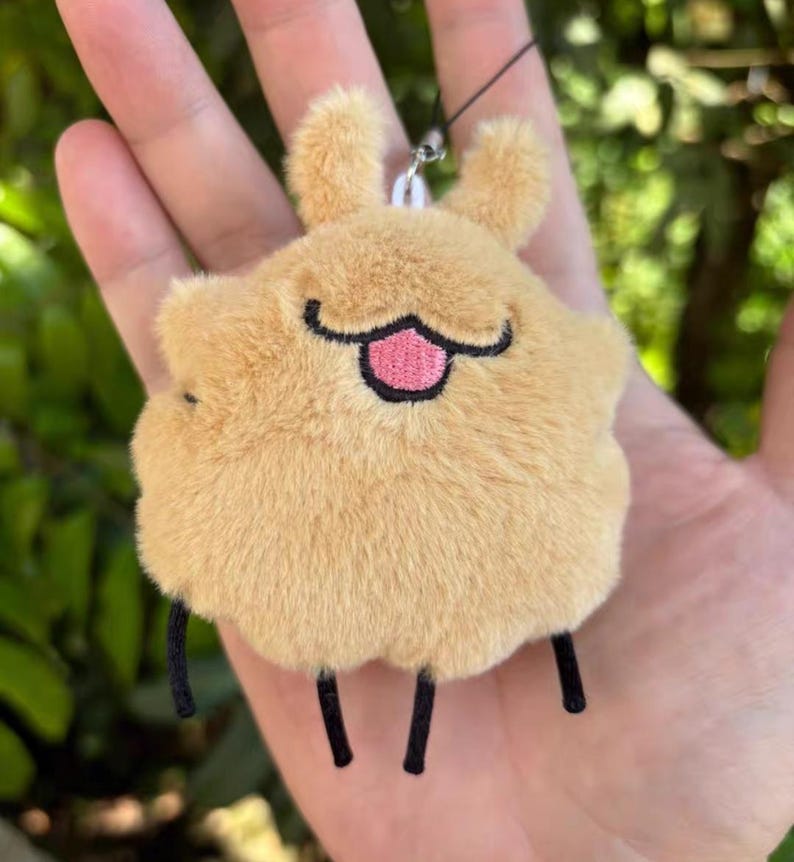 May include: A tan-coloured plush keychain in the shape of a cartoon animal. It has small, upright ears, a smiling face with a pink tongue, and three black legs. The keychain is suspended by a black string.
