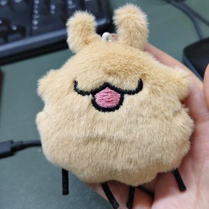 May include: A soft, tan-coloured creature with a smiling face, pink tongue, and black embroidered details. It has two small, upright tufts and black thread legs. The keychain is held in a hand.