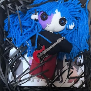 Op de afbeelding: Een handgemaakte pop met blauw garen haar, knoopogen en een paars ooglapje. De pop draagt een zwart shirt en een rode rok, met een miniatuur rode gitaar hanger. De pop wordt gepresenteerd in een zwarte doos met zwarte papieren stroken.