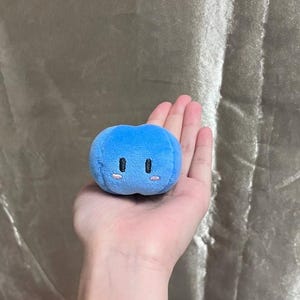 K&ouml;nnte beinhalten: Ein kleines, blaues, pl&uuml;schiges, kugelf&ouml;rmiges Spielzeug mit gestickten schwarzen Augen und rosa Wangen. Das Spielzeug wird in einer offenen Hand vor einem neutralen Hintergrund gehalten. Das Spielzeug hat eine weiche, runde Form.