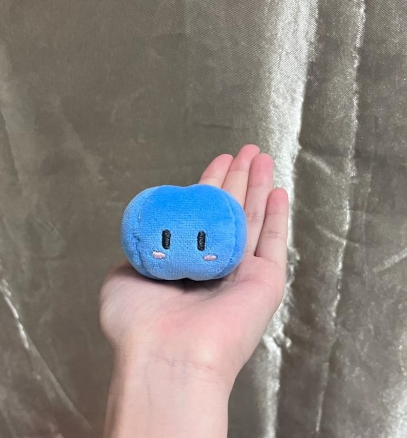 K&ouml;nnte beinhalten: Ein kleines, blaues, pl&uuml;schiges, kugelf&ouml;rmiges Spielzeug mit gestickten schwarzen Augen und rosa Wangen. Das Spielzeug wird in einer offenen Hand vor einem neutralen Hintergrund gehalten. Das Spielzeug hat eine weiche, runde Form.