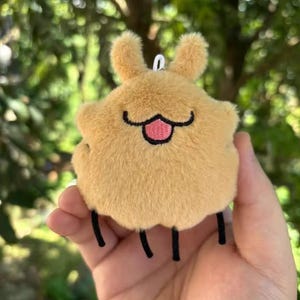May include: A tan-coloured plush keychain in the shape of a cartoon character. It features two small, fluffy ears, a black smiling mouth with a pink tongue, and three black legs. The keychain is held in a hand against a blurred green backdrop.