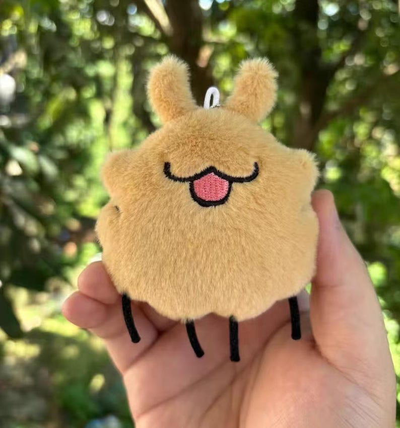May include: A tan-coloured plush keychain in the shape of a cartoon character. It features two small, fluffy ears, a black smiling mouth with a pink tongue, and three black legs. The keychain is held in a hand against a blurred green backdrop.