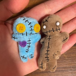 Puede incluir: Dos pequeñas muñecas de peluche hechas a mano. Una es azul claro con ojos de botón amarillos y un corazón morado. La otra es marrón con ojos de botón blancos. Ambas muñecas tienen detalles cosidos en negro.