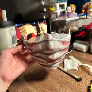 May include: A handmade face mask with a layered design. The mask features a white base with red and black accents, creating a unique, textured appearance. The mask has white ear loops and is held by a hand.