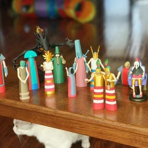 Puede incluir: Una colección de figuras coloridas y estilizadas, cada una con diseños y atuendos únicos. Las figuras están dispuestas sobre una superficie de madera, mostrando una variedad de formas y colores, incluyendo rojo, amarillo, verde y azul. Las figuras parecen hechas a mano o diseñadas a medida.