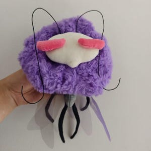 May include: A plush toy insect with a fluffy purple body, white face, and pink eye accents. It has black antennae and legs, with translucent purple wings. The toy is designed to resemble a stylized insect.