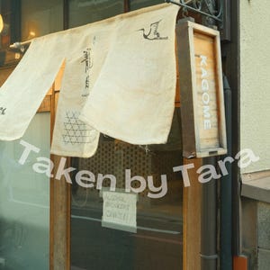 May include: Exterior shot of a Japanese restaurant entrance. A cream-colored banner with Japanese characters and a crane illustration hangs above the door. A wooden sign reads "KAGOME". A sign in the window reads "ALCOHOL DRINKERS ONLY!!"