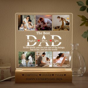 May include: A personalized illuminated acrylic sign with a collage of family photos and the text "The Best DAD" with the names "Chrystal", "Oliver", and "Tyler" and the message "To the world, you are just one of the beings; but to me, you are the world". The base of the sign is wood with the text "❤CHRYSTAL OLIVER TYLER❤ HAPPY FATHER'S DAY 2024"