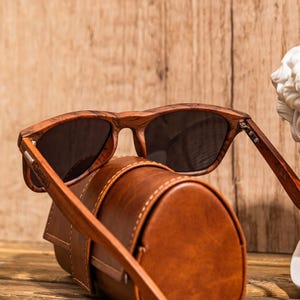 Puede incluir: Gafas de sol de madera marr&oacute;n con lentes oscuros y una funda de cuero marr&oacute;n. Las gafas tienen la palabra "Gerald" grabada en el lateral.