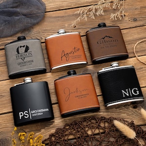 Puede incluir: Seis petacas personalizadas con varios diseños y textos. Las petacas son de diferentes colores, incluyendo negro, marrón y gris, con detalles plateados. Cada petaca tiene una inscripción única, como nombres, fechas y títulos.