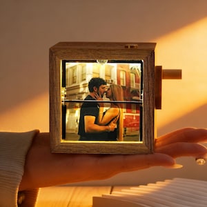 以下が含まれることがあります： 温かい光で照らされた、キスをしているカップルの写真が入った木製のフォトフレーム。 フレームは長方形で、明るい茶色の仕上げです。 小さな木製のハンドルが側面に付いています。